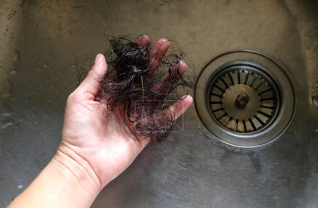 Photo for Hand pulls hair out of sink in kitchen drain. Female palm extracts shock of hair from siphon in  sink in kitchen. Water drain hole clogged with hair clump. Close up. Cleaning - Royalty Free Image