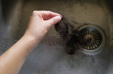 Hand pulls hair out of sink in kitchen drain. Female palm extracts shock of hair from siphon in  sink in kitchen. Water drain hole clogged with hair clump. Close up. Cleaning
