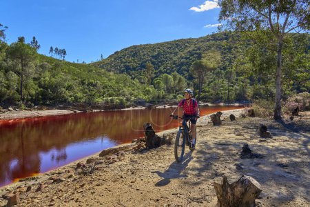 Téléchargez les photos : Belle femme avec vélo de montagne électrique lors d'un tour à vélo le long de la rivière Rio Tinto avec son eau rouge naturelle en Andalousie, Espagne - en image libre de droit