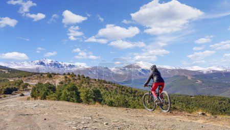 Foto de Agradable, activa mujer mayor ciclismo con su bicicleta de montaña eléctrica por debajo de las montañas cubiertas de nieve de la Sierra Nevada española, cerca de Granada, Anslusia, España - Imagen libre de derechos
