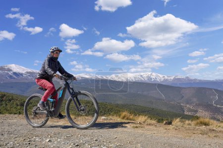 Foto de Agradable, activa mujer mayor ciclismo con su bicicleta de montaña eléctrica por debajo de las montañas cubiertas de nieve de la Sierra Nevada española, cerca de Granada, Anslusia, España - Imagen libre de derechos