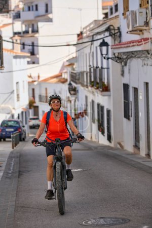 Nette Seniorin radelt mit ihrem Elektro-Mountainbike im malerischen weißen Dorf Frigiliana in der Nähe von Nerja, Andalusien, Spanien