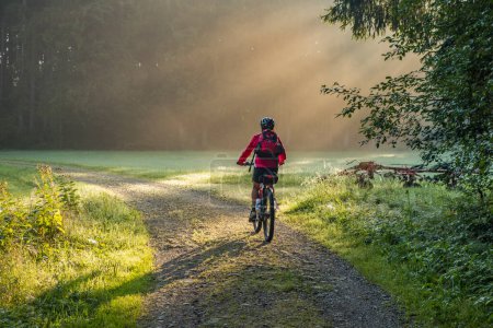 Téléchargez les photos : Femme âgée, chevauchant un vélo de montagne électrique par une matinée atmosphérique au lever du soleil - en image libre de droit