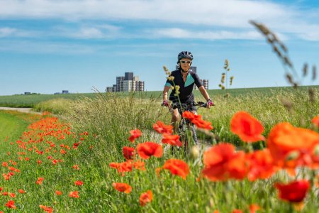 Foto de Mujer mayor agradable, montar una e-Mountainbike en el suburbio de una gran ciudad, rodeado de campos verdes y amapolas rojas - Imagen libre de derechos