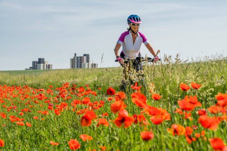 Photo for Nice senior woman, riding a e-Mountainbike in the suburb of a big city, surrounded by green fields and red poppies - Royalty Free Image