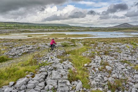 Photo for Nice senior woman on mountain bike, cycling in the rough karst area of Burren near Ballyvaughan, County Clare in the western part of the Republic of Ireland - Royalty Free Image