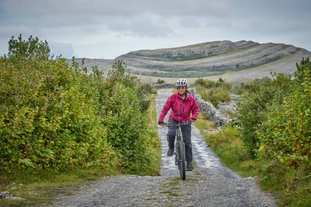 agradable mujer mayor en bicicleta de montaña, ciclismo en la áspera zona kárstica de Burren cerca de Ballyvaughan, Condado de Clare en la parte occidental de la República de Irlanda