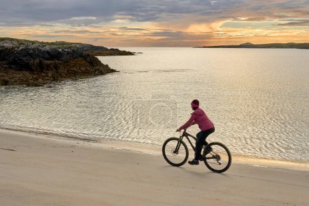 Téléchargez les photos : Belle femme âgée en VTT, vélo au coucher du soleil sur la plage de sable doré de Sillerna, Grallagh, comté de Galway, dans la partie ouest de la République d'Irlande - en image libre de droit