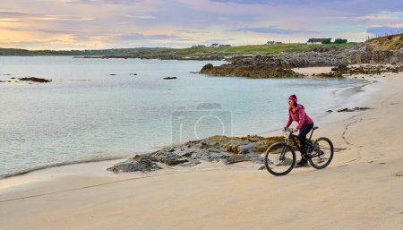 Téléchargez les photos : Belle femme âgée en VTT, vélo au coucher du soleil sur la plage de sable doré de Sillerna, Grallagh, comté de Galway, dans la partie ouest de la République d'Irlande - en image libre de droit