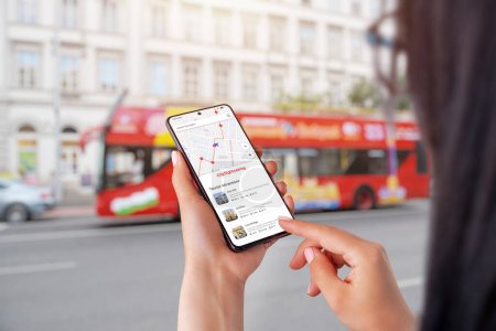 City Sightseeing app on smartphone in woman hands. City tourist bus in background
