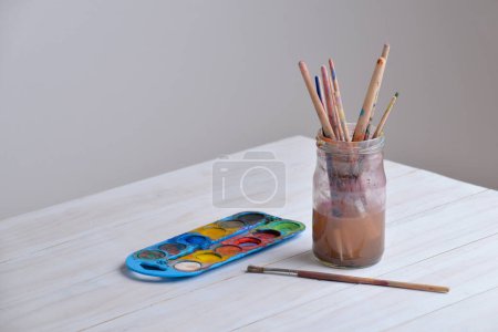 Jar with paintbrushes on work table with watercolors. Copy space aside. Ideal for art supplies, creativity, or painting concepts