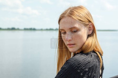 schöne Mädchen in einem schwarzen Kleid auf dem Hintergrund des Flusses, Meer mit blauem Himmel und Wolken Nahaufnahme
