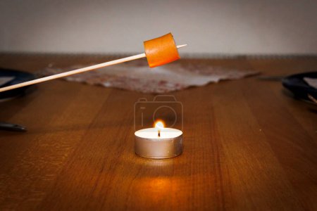Foto de Un trozo de salchicha se asa en un pincho de madera sobre la llama de una luz de té colocada en una mesa de madera de haya - Imagen libre de derechos