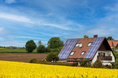 Single-family house with photovoltaic modules on the roof in front of a rape field in full bloom under a blue sky