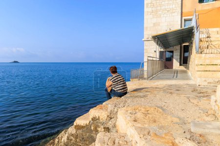 Téléchargez les photos : Femme caucasienne âgée avec les cheveux courts de derrière se trouve sur les marches du mur du port de la ville croate Rovinj et regarde vers la mer à petite île - en image libre de droit