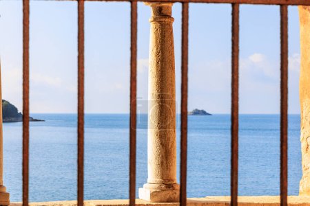 Foto de Vista a través de una ventana con barras de una columna erosionada frente al mar en la ciudad de Rovinj en Croacia en un día de verano con cielo azul - Imagen libre de derechos