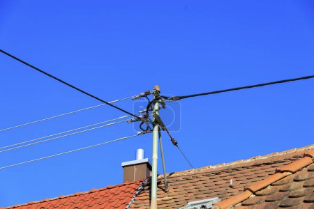 Téléchargez les photos : Mât électrique sur une maison couverte de tuiles de toit devant une cheminée sous le ciel bleu - en image libre de droit