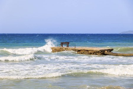 Téléchargez les photos : Un banc de repos au milieu des vagues sur l'île de Corfou près de la ville d'Arillas sous un ciel bleu et une mer lourde - en image libre de droit