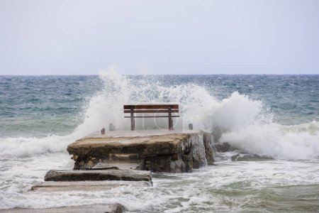 Foto de Un banco de descanso en medio de las olas en la isla de Corfú cerca de la ciudad de Arillas bajo cielos azules y mares pesados - Imagen libre de derechos
