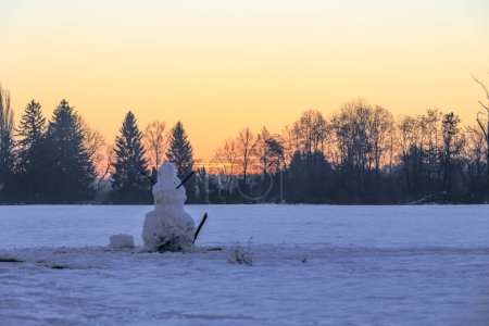 Téléchargez les photos : Un grand bonhomme de neige se tient au lever du soleil sur une prairie enneigée à Siebenbrunn près d'Augsbourg contre un ciel coloré avec des arbres à l'horizon - en image libre de droit