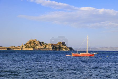 Téléchargez les photos : Un voilier rouge ancre dans la mer bleue en face de la forteresse de Corfou ville à la lumière du soleil du soir sous un ciel bleu avec des nuages légers - en image libre de droit