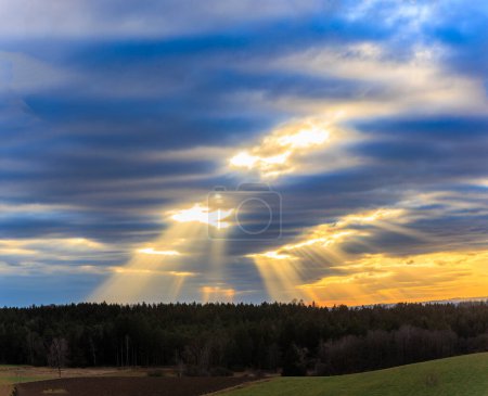 Sonnenstrahlen brechen an einem bewölkten Wintertag durch eine dichte Wolkendecke über einer Waldlandschaft bei Ravensburg in Baden-Württemberg.