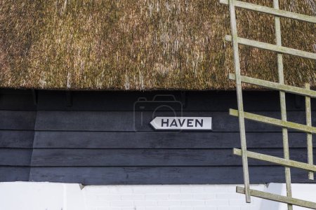 Un letrero con el texto holandés Puerto en una pared de madera pintada oscura bajo un techo de paja de un molino de viento de pólder para drenar las zanjas en una zona residencial en Den Oever en los Países Bajos, 24.3.2024