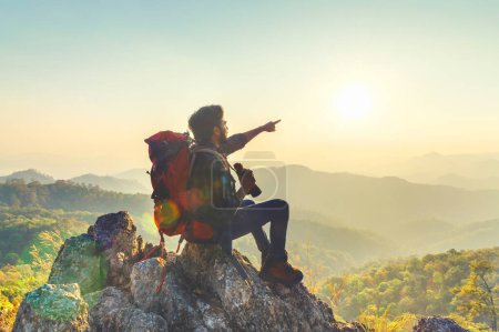 Téléchargez les photos : Voyageur avec sac à dos et tenant jumelle assis sur le sommet de la montagne pointant vers la vue - en image libre de droit