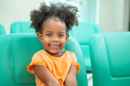 Téléchargez les photos : Douce petite fille à l'extérieur avec les cheveux bouclés souriant assis sur la chaise à la chambre - en image libre de droit