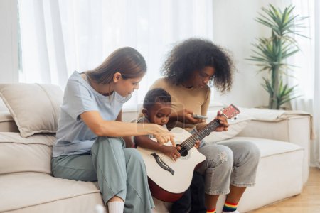 Téléchargez les photos : Famille LGBT diversité lesbienne couple avec son garçon apprendre la guitare dans la chambre. Mode de vie LGBT - en image libre de droit