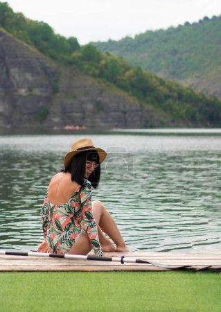 Téléchargez les photos : Une fille en maillot de bain avec des manches est assise sur la jetée. une rame se trouve à proximité. été près de la rivière. photo esthétique. rochers et rivière. Photo de haute qualité - en image libre de droit