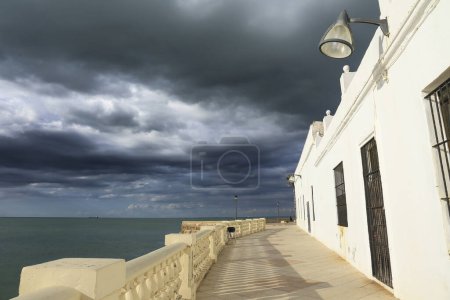 Téléchargez les photos : Maisons blanchies à la chaux dans la promenade de la plage de Chipiona par une journée orageuse - en image libre de droit