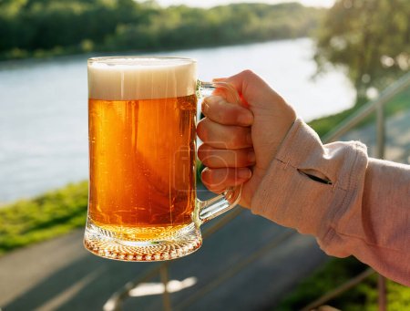 A hand holding a glass mug filled with golden beer, with a scenic river and lush green landscape in the background. The beer looks refreshing with its foamy head and the bright sunlight reflecting off the glass.