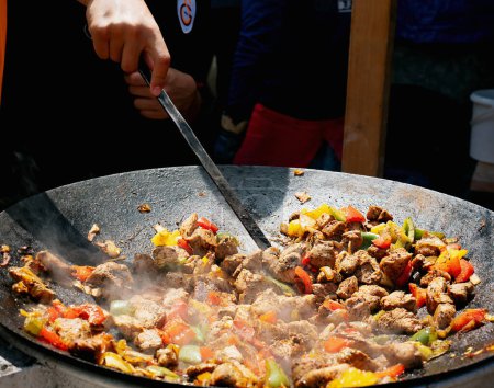 Hand stirring seasoned meat and colorful bell peppers in a large cast-iron skillet, with steam rising from the hot pan.
