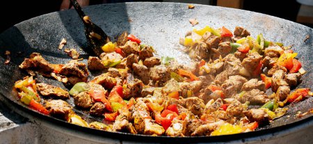 Hand stirring seasoned meat and colorful bell peppers in a large cast-iron skillet, with steam rising from the hot pan.