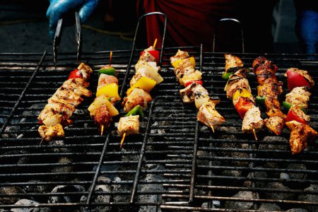 Person in gloves turning skewers of marinated meat and vegetables on a hot grill, with smoke rising.