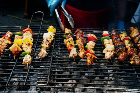 Person in gloves turning skewers of marinated meat and vegetables on a hot grill, with smoke rising.