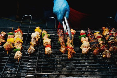 Foto de Persona con guantes haciendo brochetas de carne y verduras marinadas en una parrilla caliente, con humo subiendo. - Imagen libre de derechos