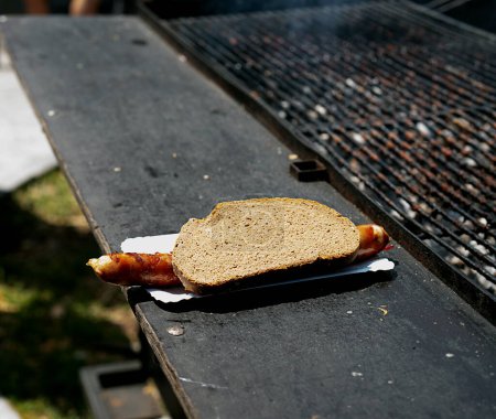A grilled sausage served in a slice of bread, placed on a paper plate, resting on a barbecue grill table.