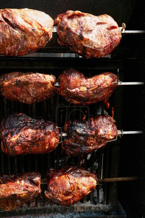 Multiple cuts of meat on a rotisserie grill, with flames and smoke, highlighting the rustic cooking process.