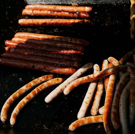 A variety of sausages grilling on a hot barbecue, showcasing both white sausage and kaiserkraner sausages with grill marks.