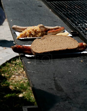 Grilled sausage in a slice of bread on a paper plate, with additional sausages and sauerkraut on the grill table.