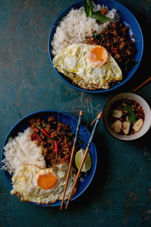 Two blue plates with Pad Kra Pow, featuring stir-fried minced pork, fried egg, steamed rice, and lime. Bowl of chili garlic sauce. Wooden chopsticks. Fresh basil garnish. Overhead view on textured blue background.