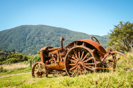 Foto de Tractor agrícola vintage desertó y se oxidó en el claro en el campo de arbustos y colinas de South Island. - Imagen libre de derechos