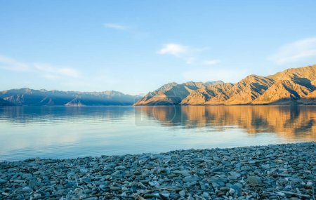 Photo for Stony foreground of peaceful Lake Wanaka in morning light surrounding hills and blue sky. - Royalty Free Image