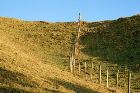 Photo for Fence leading up and over slope under blue sky in agricultural background. - Royalty Free Image
