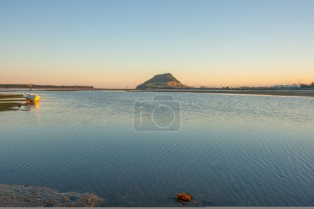 Foto de Monumento Monte Maunganui en el horizonte distante al amanecer con el bote clinker pasado de moda en la playa en Harbour Drive Tauranga. - Imagen libre de derechos