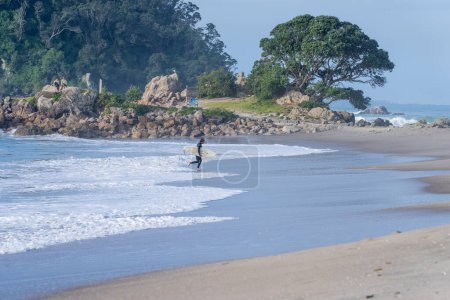 Foto de Tauranga Nueva Zelanda - 26 de junio de 2023; Surfista en traje de neopreno que transporta tablas de surf desde el mar hasta la playa. - Imagen libre de derechos