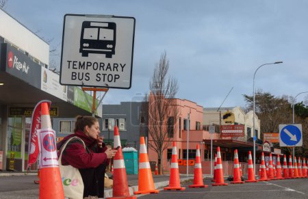Téléchargez les photos : Tauranga Nouvelle-Zélande - 31 juillet 2023 ; Les cônes de route dominent le paysage de la rue tandis que la femme s'assoit sur le trottoir sous un panneau d'autobus temporaire fumant et regardant appareil mobile - en image libre de droit