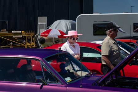Foto de Mount Maunganui Nueva Zelanda - 12 de noviembre de 2023; All USA Car Day - Mujer con gafas de sol en forma de corazón y sombrero rosa camina entre los coches. - Imagen libre de derechos